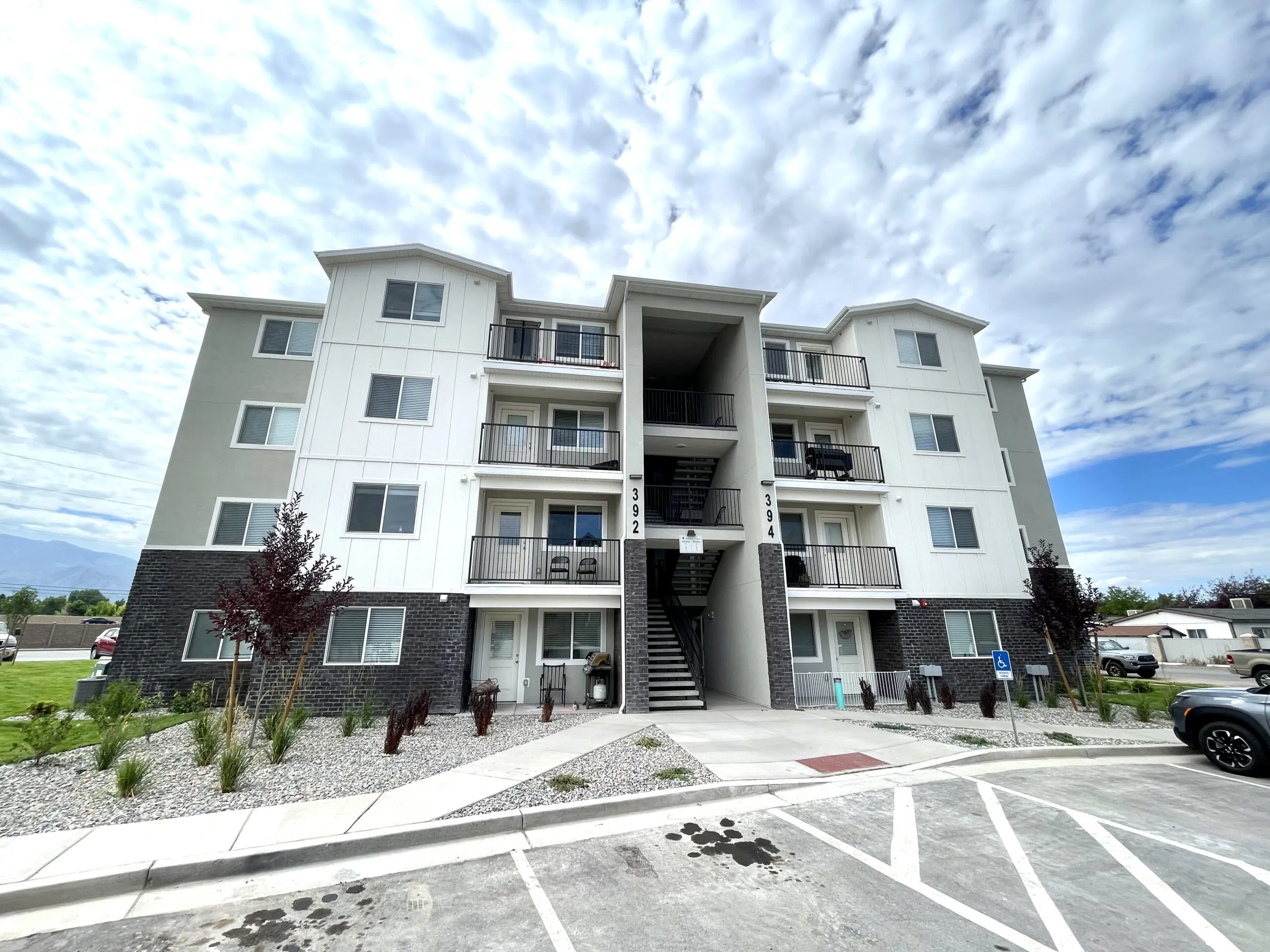 Four-story modern apartment building with balconies and central staircase. The facade is gray and white, with a landscaped front. Cloudy sky backdrop.