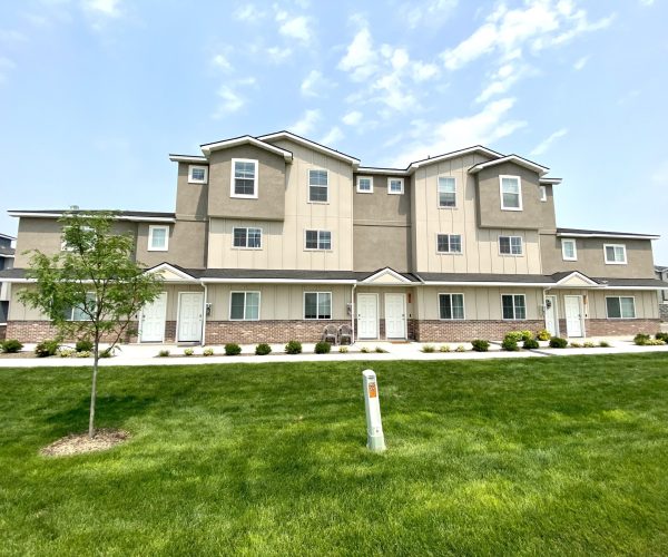New Home Construction 3 Three-story beige townhouse with multiple windows and white doors, surrounded by a well-maintained green lawn and a small tree under a blue sky.
